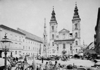 Piac a belv�rosi pl�b�niatemplomn�l. A templom el�tt a Szenth�roms�g-szobor �ll, balra a kegyesrendiek rendh�za emelkedik. 1877. Kl�sz Gy�rgy felv�tele. Budapesti T�rt�neti M�zeum